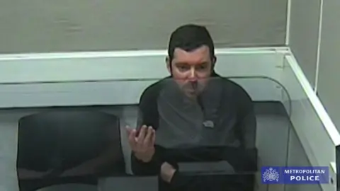 Metropolitan Police Scott O'Connor in a police interview. He is wearing a black top and sitting behind a transparent screen in a room with grey walls. 