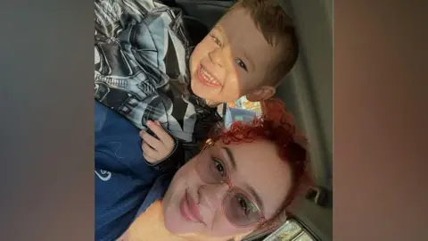 Courtney A woman with pink rimmed glasses and clurly red hair pulled back is in a car and on her right shoulder a young boy in a batman graphic top is smiling