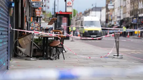 PA Media An exterior view of the restaurant where the victims were attacked in Dalston. There is a police tape cordon in place, and a police van blurred in the distance, There is debris on the pavement.