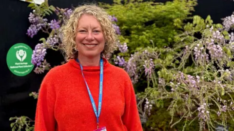 Simone Daye smiling into the camera in front of purple wisteria. She has short, blonde curly hair and is wearing a bright red jumper and blue lanyard.