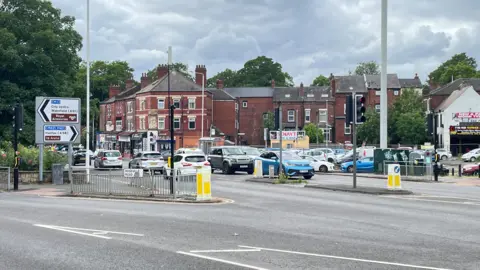 Several cars travelling in both directions at a road junction. Some of them are waiting at a set of traffic lights. The main road has a section which goes off to the left.