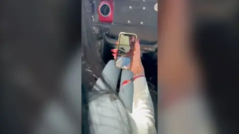 A woman inside a car holds a smartphone to video call a young man.