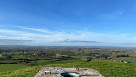 The view from the top of Cley Hill, with some of the top in the foreground and a concrete marker. Beyond, green fields and hedges stretch into the distance under a blue sky.