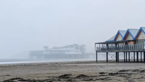 Gary the Monkey/Weather Watchers Weston-super-Mare's pier is disappearing behind fog. The beach is clear in the foreground but fog is rising above the water in the background.