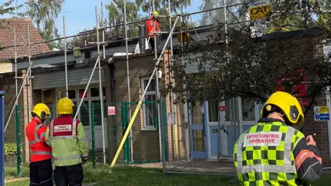 Three firefighters with their backs to the viewer looking at a low-lying school building with scaffolding around it after a fire