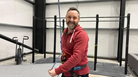 Alan is wearing a harness with a rope attached, belaying Sam who is off camera. Alan is wearing a red hoodie and is smiling at the camera.