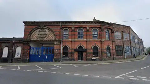 A red brick building which has large blue gates with an ornate archway above them. The building is on the corner of a road junction.