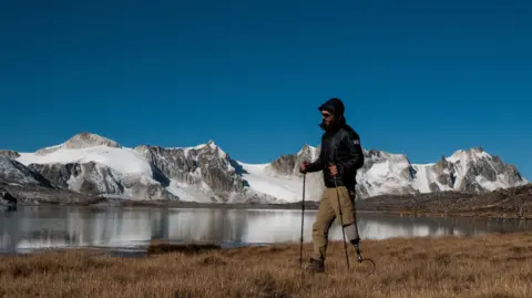 Rich Potter/Rich Davies Rich Potter, who is wearing a black-hooded coat, sunglasses, and light brown trousers, walking in front of snow-covered hills and a lake. He is wearing a black boot on his left foot and has an artificial right leg. 