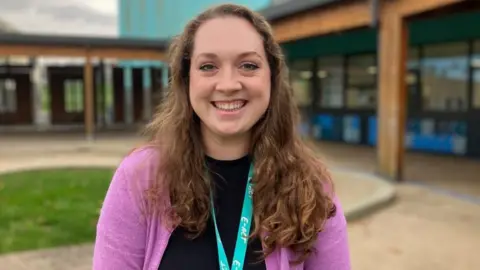 Hannah Green. She is smiling at the camera, and has long brown hair, and is wearing a purple top