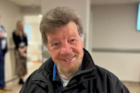 Ronald Muir has greying hair and a clipped beard and is pictured in the waiting room of the urgent access centre in Carlisle. He is smiling and wearing a black waterproof jacket.