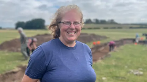 A woman with blonde hair and glasses, wearing a blue t-shirt, looks at the camera