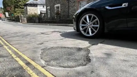 A pothole on Dolvin Road in Tavistock with a passing navy blue car.