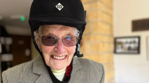 Elizabeth is looking directly at the camera and smiling. She is wearing a black riding hat, tinted glasses, a cream polo neck top with tweed jacket. The photo is a close up of her head and shoulders. 
