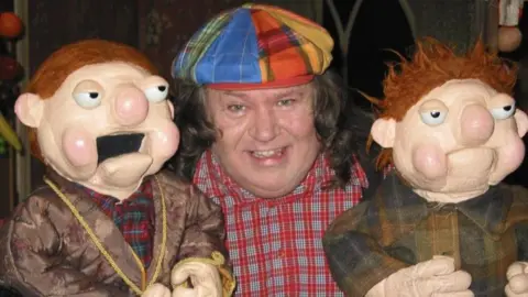 Richie Kavanagh A man smiling at the camera, he has long brown hair and a multi colour flat cap on. He is wearing a red and white check shirt. Either side him of his a puppet of a man that has red hair.