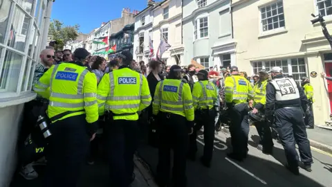 Claudia Sermbezis Police officers in yellow hi-vis standing in front of a crowd of people, in a narrow street. 