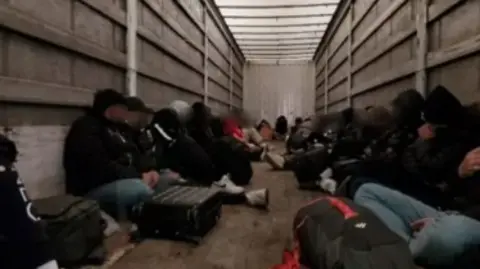 Dozens of people are seated on the floor inside a large lorry. Their faces are blurred in the image and many appear to be male in jeans and hooded tops and jackets.
