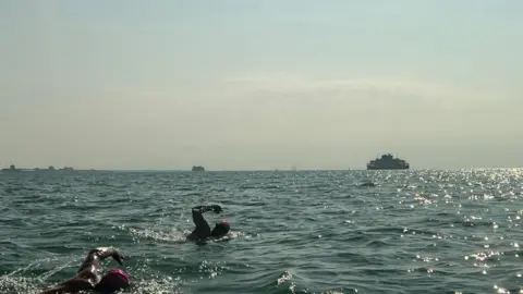 The two men swimming in the sea, with ferries visible on the horizon