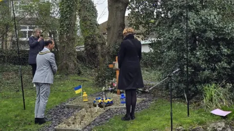 Ewhen Chymera Two women and a man stand in front of a cross on the floor with blue and yellow flags and flowers