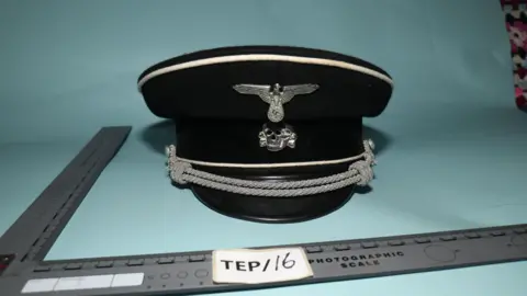 PA Media A counter-terror police image of a black and silver peaked military cap with a silver skull and Nazi Swastika emblem on the front. There is a right-angled ruler to show scale.