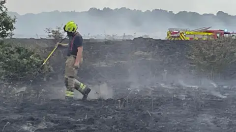 A single firefighter walking around a blackened field with wisps of smoke coming off it and a fire engine in the background