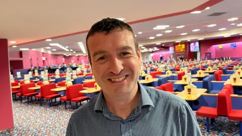 John Devine/BBC Dan Shuttleworth, 48, has short dark hair and is smiling, he is wearing a black and white striped shirt and behind him is a bingo hall with lots of red and blue chairs around small tables for marking off numbers, there are large screens at the back displaying the numbers.