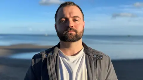 Devon Watson has short dark hair and a short dark beard. He is wearing a white t-shirt under a grey jacket. He is standing in front of a blurred out beach on a sunny and slightly cloudy day.