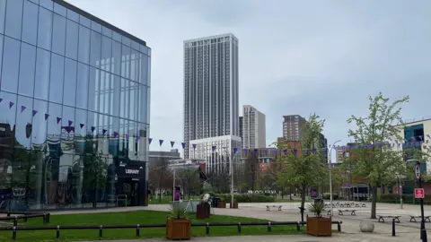 A view across a paved public square at Aston University, with the campus library on the left and purple bunting strung across the space. In the distance the tall white One Eastside tower rises above other city centre buildings under a grey sky.