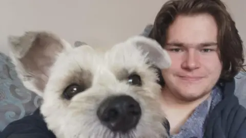 Leigh Day Solicitors A man in his early 20s, with long brown hair and brown eyes sits on a turquoise patterned sofa. A small white terrier type dog is sitting on his lap with its face close to the camera lens. 