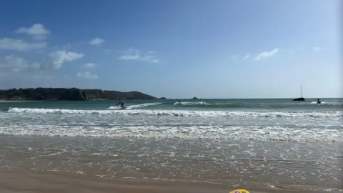People in the water at St Brelade Bay. It is a sandy beach and there's a view of waves crashing on the sand. There's land in the distance on the left. The sky is blue with a few white clouds.
