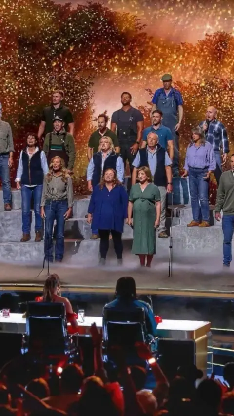The Hawkstone Farmers Choir performs on the Britain’s Got Talent stage in front of the judges and a packed audience. Bathed in golden lights and sparkling effects, the choir delivers a powerful performance that celebrates rural life and community spirit. Standing on tiered platforms, the singers are led by their conductor as they captivate the crowd with an emotional and uplifting rendition.
