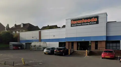 Google Un gran edificio gris con las palabras Farm Foods en letras rojas y un estacionamiento en frente.