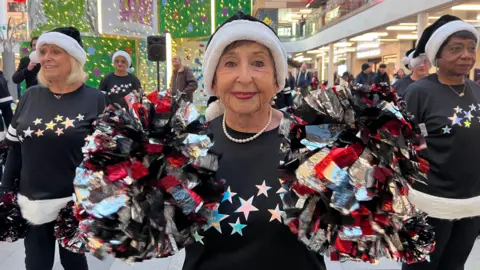  A close-up of a performer in a shopping centre holding two large pom-poms in red, black, silver, and white. The person is wearing a black sweater with colourful star designs across the chest and a festive hat with white trim. 