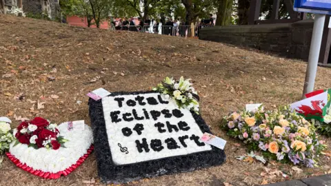 A white and red heart-shaped floral tribute is laid on the ground next to a black and white 'Total Eclipse of the Heart' tribute, alongside a tribute of lilac, pink and peach flowers. A Welsh flag is also laid on the ground nearby alongside a Portugese flag. 