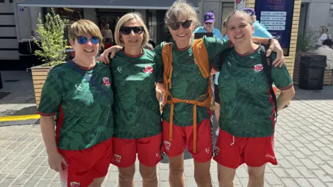 Four members of the Wales women's walking football squad. They are wearing red shorts and green football shirts with the Welsh flag on, and have their arms around each other, smiling at the camera. Three of the women are wearing sunglasses. They are stood on a street in the sunshine.