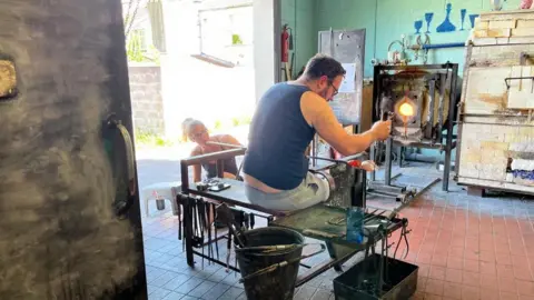 A man and woman work in a factory setting, shaping glass, using artisanal tools, with a burning hot furnace in the background. The woman on the left, is on her knees, using a glass-blowing funnel, while a man on the left, is carefully shaping with specialised tools. 