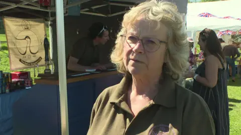 A woman looking to the left of the camera. She is standing outdoors next to a pop-up stall at a fete. 
