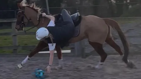 Danielle Pratt A young woman hanging from a horse's back to the floor to pick up a ball. She has a long blonde plait and is wearing black jodhpurs, a black T-shirt with white sleeves and a black helmet. The horse is chestnut with white socks and is cantering around a sandy ring.