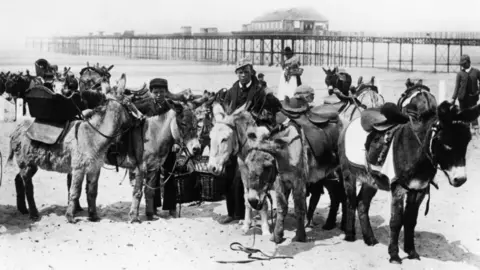 Getty Images A group of donkeys on the beach in Rhyl with saddles and bridles on and a handler standing in the middle of them. The picture is black and white and a man and woman in 1890s clothing can be seen in the background, as can the pier.