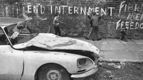 Brendan Murphy A black and white image of 5 young boys. One is sitting in a car with his head out the window, while another points a fake gun towards a boy who is holding another up against a wall. Then there is a boy on his knees sitting beside them.