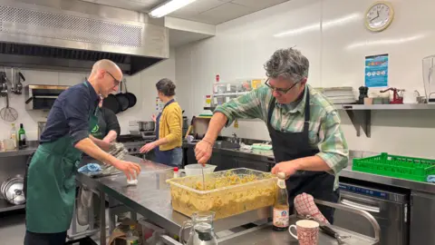 A group of people wearing aprons preparing food around stainless-steel workstations in a kitchen.
