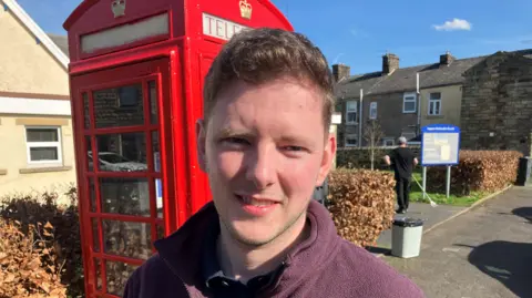 An image of councillor Jamie McGowan standing in front of a red phone box. He is smiling and wearing a purple fleece. In the background a man is gardening next to a sign for the church.