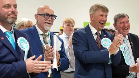 four male Reform candidates with rosettes laugh and clap 