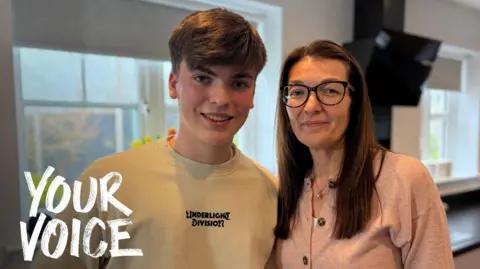 A man and his mum look at the camera - both are smiling. It is a head and shoulders shot of the two of them. The man has a cream jumper on, while the woman has a pink shirt.