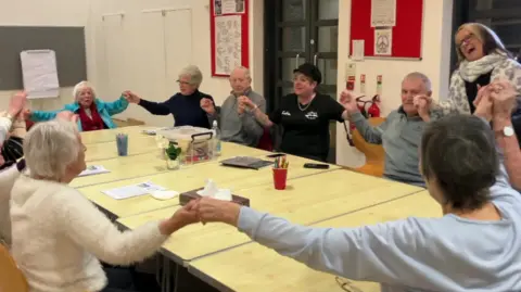 Shot of a group of mostly elderly people, sat holding hands and singing together in a circle 