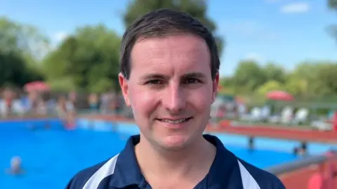 Matt Day is smiling at the camera. He has short, dark hair and is clean-shaven. He is wearing a dark blue polo shirt with white stripes across the shoulders. Behind him is a blurred image of a large swimming pool with sunbeds and parasols