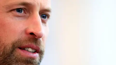 A close-up of Prince William's face. He looks like he is concentrating on something as his eyes are focussed. He has a brown and grey-flecked beard and moustache and blue eyes.  