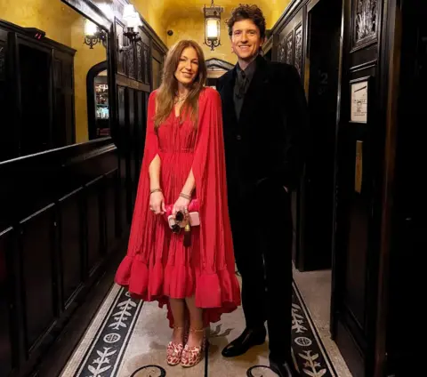 Greg James Bella wears a red long dress with cape and has long brown hair. Greg James wears a dark coloured suit with a grey tie and has short brown hair
