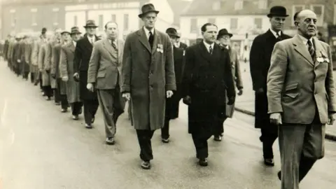Swaffham Museum A sepia-toned black and white image of men marching through the streets of Swaffham in 1959. They are wearing suits, some with overcoat. Many have hats on and have medals pinned to their left breast.