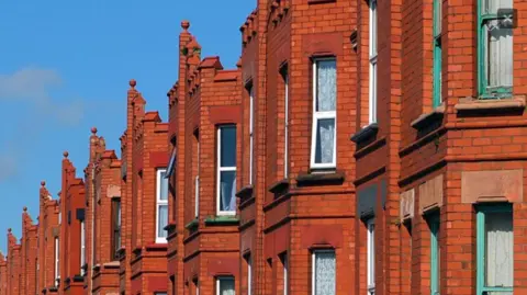 A row of red-bricked buildings with rectangular windows, with a blue sky in the background.