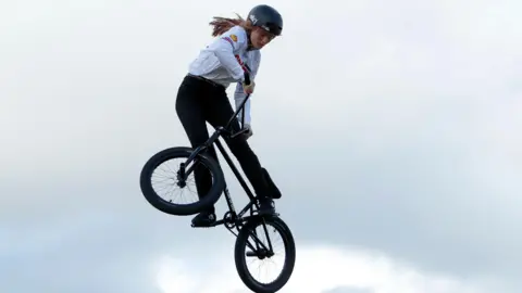 A woman wearing a black helmet, white top and black trousers, is mid-air while she performs a trick on a BMX.